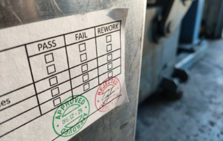 Witte inspectielabel met zwarte vakjes en groene en rode stempels op industrieel metalen oppervlak