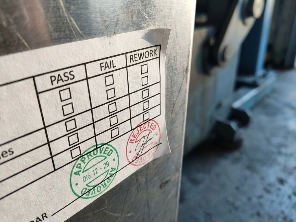 inspectie-label-kwaliteitscontrole-industrieel.jpg Witte inspectielabel met zwarte vakjes en groene en rode stempels op industrieel metalen oppervlak